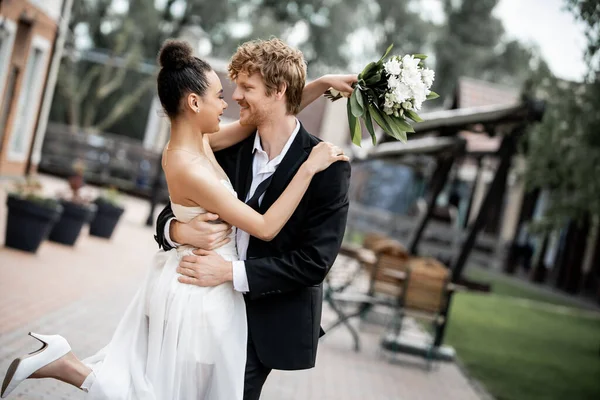 Matrimonio in città, sposa afro-americana con fiori e felice elegante sposo che abbraccia per strada — Foto stock
