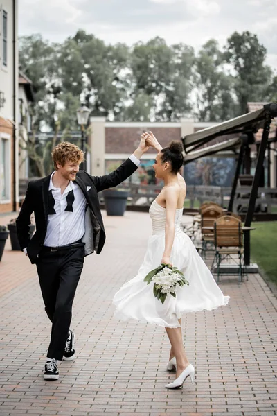 Свадьба в современном городе, элегантная многонациональная пара, держащаяся за руки и танцующая на улице, счастье — стоковое фото