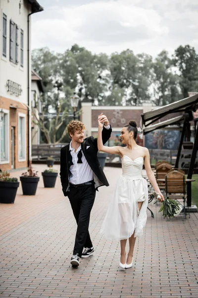 Alegres recién casados multiétnicos bailando y tomados de la mano en la calle de la ciudad, celebración al aire libre - foto de stock