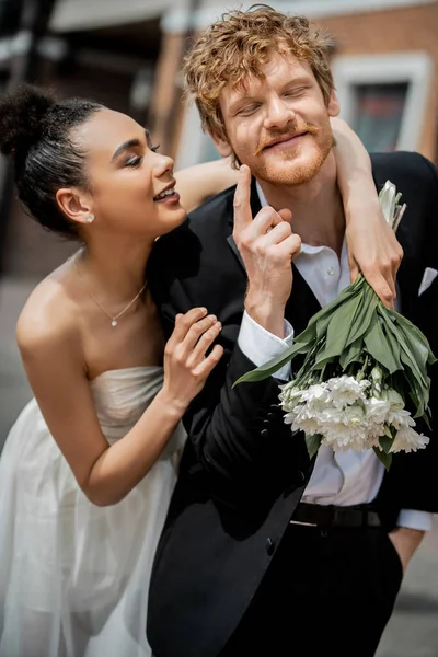 Homem ruiva feliz apontando para bochecha perto de noiva americana africana feliz com flores, casamento ao ar livre — Fotografia de Stock