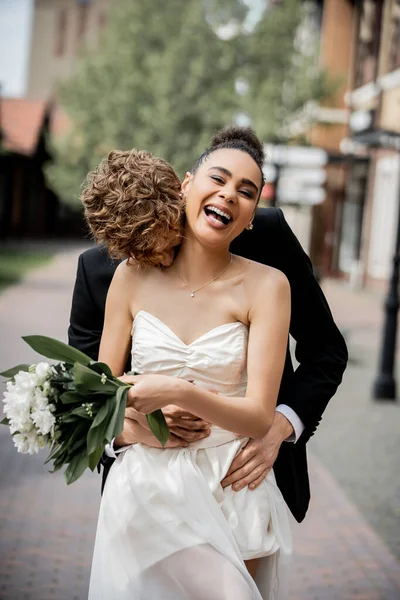 Rossa sposo abbracciare ridere africano americano sposa con bouquet, festa di nozze in città — Foto stock