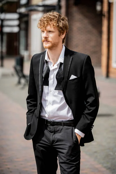 Elegant redhead man in black dress standing with hands in pockets on street, groom, outdoor wedding — Stock Photo