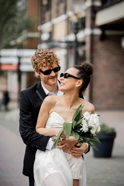 Casal interracial com hambúrgueres e suco de laranja perto da fonte da cidade, traje de casamento, óculos de sol — Fotografia de Stock
