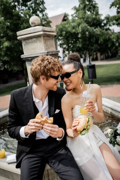 Sposi multietnici che celebrano il matrimonio vicino alla fontana della città, hamburger, succo d'arancia, spuntino — Foto stock