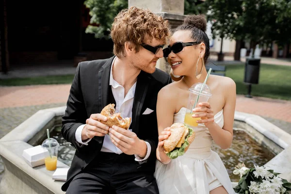 Elegant interracial newlyweds in sunglasses with burgers and orange juice near city fountain — Stock Photo