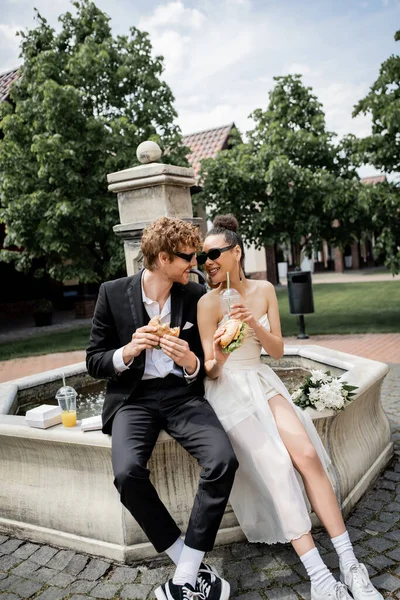 Heureux couple interracial en tenue de mariage et lunettes de soleil avec hamburgers et jus près de la fontaine de la ville — Photo de stock