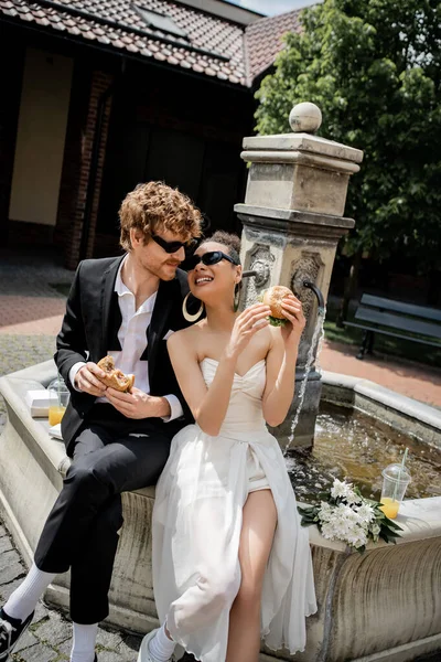 Recién casados multiétnicos en gafas de sol snack con hamburguesas y zumo de naranja cerca de la fuente de la ciudad - foto de stock