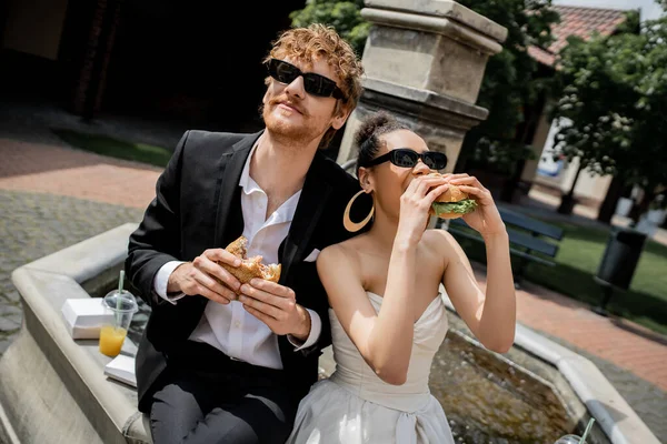 Afrikanisch-amerikanische Braut isst Burger in der Nähe von rothaarigen Bräutigam in Sonnenbrille in der Nähe von Brunnen, Outdoor-Hochzeit — Stockfoto