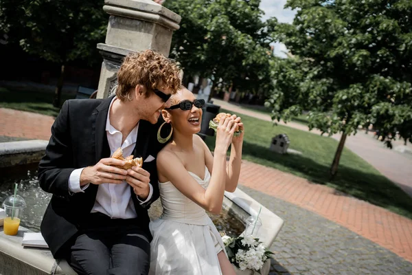 Animado casal multiétnico comemorando casamento perto da fonte da cidade, óculos de sol, hambúrgueres, suco de laranja — Fotografia de Stock
