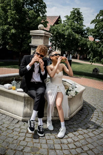 Multiethnic newlyweds in sunglasses eating burgers near fountain, wedding in european city — Stock Photo