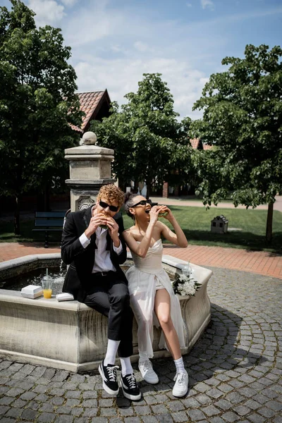Элегантные межрасовые молодожены в солнечных очках едят бургеры возле фонтана в европейском городе — стоковое фото