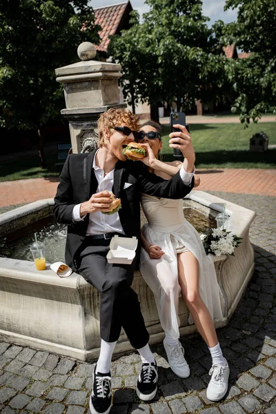 Mulher americana africana com hambúrguer alimentando noivo tomando selfie no smartphone perto da fonte da cidade — Fotografia de Stock