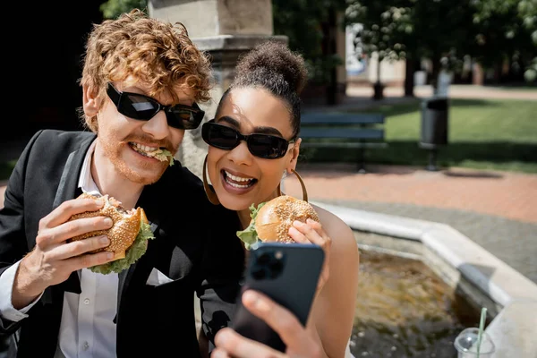 Recién casados interracial en gafas de sol sosteniendo hamburguesas y tomando selfie en el teléfono inteligente en la ciudad - foto de stock