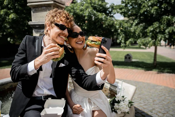 Couple multiethnique en tenue de mariage et lunettes de soleil serpentant avec des hamburgers et prendre selfie en ville — Photo de stock