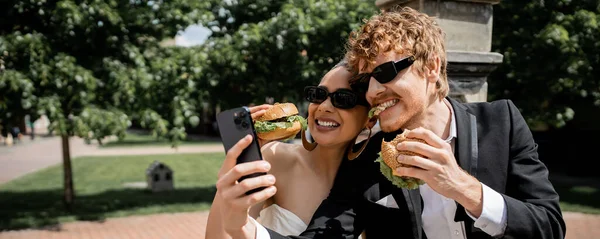 Interracial newlyweds in sunglasses eating burgers and taking selfie on smartphone on street, banner — Stock Photo