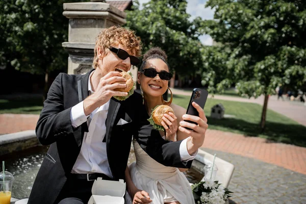 Multiethnic newlywed couple in sunglasses eating burgers, taking selfie on smartphone near fountain — Stock Photo