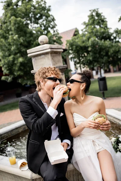 Pareja interracial en traje de novia y gafas de sol comiendo hamburguesa juntos cerca de la fuente en la ciudad - foto de stock