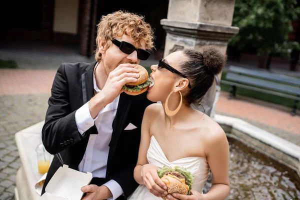 Interracial recién casados en gafas de sol mordiendo divertirse y mordiendo hamburguesa juntos cerca de la fuente - foto de stock