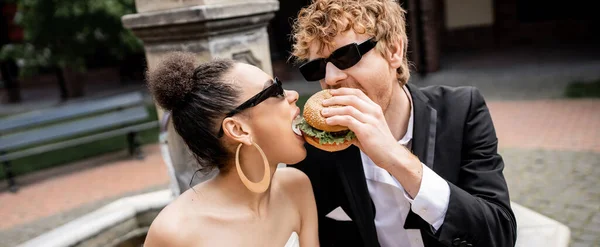 Multiethnic newlywed couple in sunglasses biting burger together, having fun outdoors, banner — Stock Photo