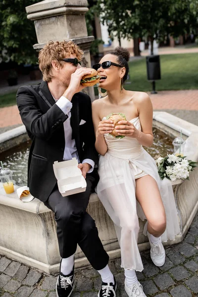 Elegante interracial recém-casados em óculos de sol comer hambúrguer juntos, rua da cidade, fonte, diversão — Fotografia de Stock