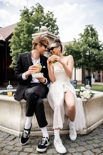Joyful interracial newlyweds in sunglasses holding burgers and looking at each other near fountain — Stock Photo