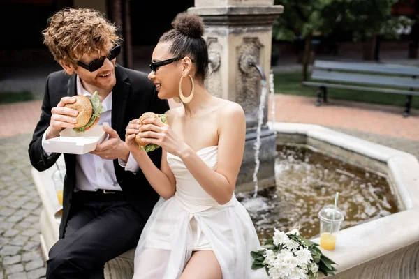 Outdoor wedding, interracial couple in sunglasses snacking with burgers at fountain in european city — Stock Photo