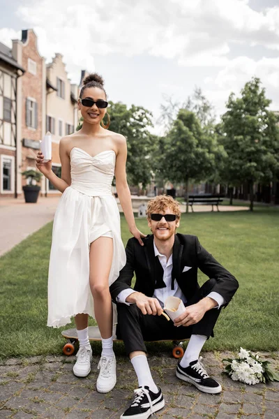 Feliz casal interracial, traje de casamento, óculos de sol, com batatas fritas, olhando para a câmera na cidade — Fotografia de Stock