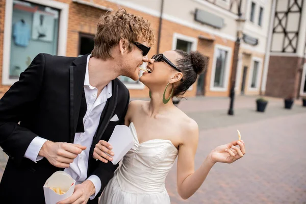 Romance urbano, boda al aire libre, recién casados multiétnicos en gafas de sol comiendo papas fritas juntos - foto de stock