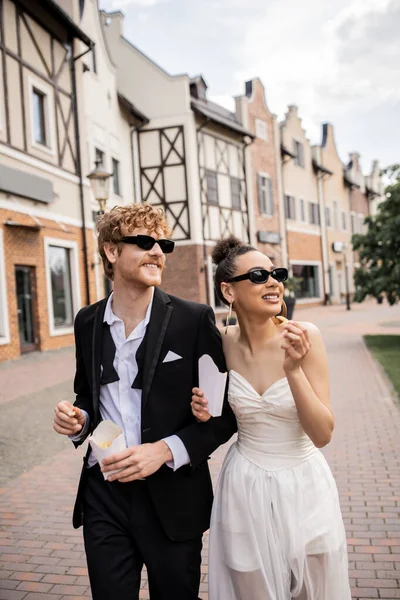 Elegante casal multiétnico com batatas fritas andando na cidade, traje de casamento, óculos de sol, felicidade — Fotografia de Stock