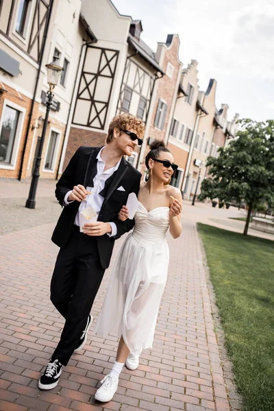 Recién casados multiétnicos con papas fritas caminando por la calle, gafas de sol, boda en un entorno urbano - foto de stock