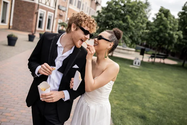 Felice sposa afro-americana che alimenta lo sposo elegante con patatine fritte, matrimonio in strada — Foto stock
