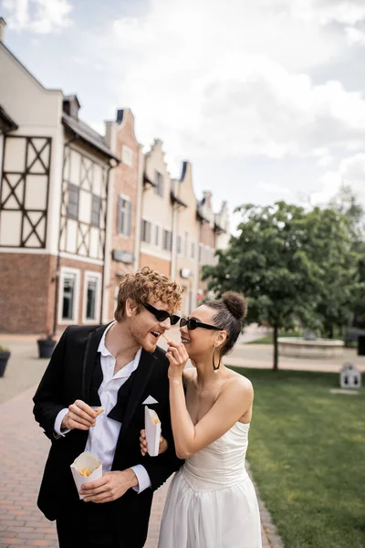 Matrimonio in città, divertimento, sposa afro-americana in occhiali da sole che alimenta lo sposo rossa con patatine fritte — Foto stock