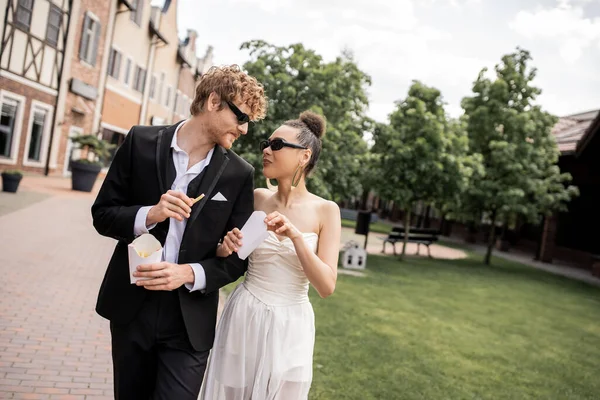 Interracial newlywed couple in sunglasses, with french fries, looking at each other on street — Stock Photo