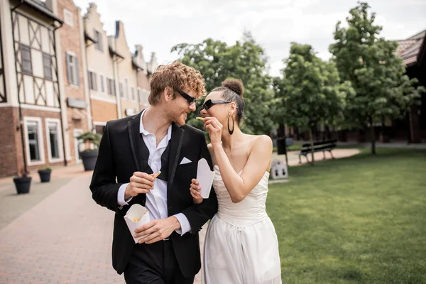 Elegante sposa afroamericana che alimenta lo sposo rossa con patatine fritte, occhiali da sole, strada urbana — Foto stock