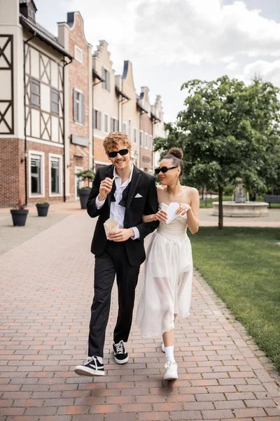 Ruiva homem e mulher americana africana andando com batatas fritas na cidade, traje de casamento, óculos de sol — Fotografia de Stock