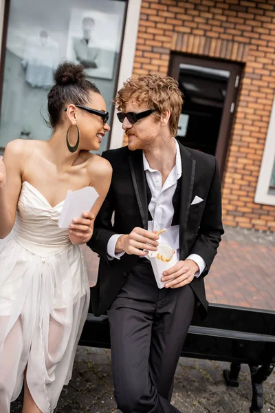 Веселая многонациональная пара в солнечных очках, с картошкой фри, смотрящая друг на друга на городской улице — стоковое фото
