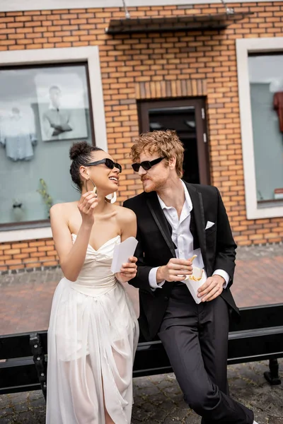 Novia afroamericana con papas fritas sonriendo cerca elegante pelirroja novio, boda en la ciudad moderna - foto de stock