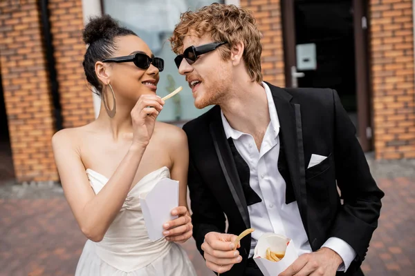 Boda en la ciudad, diversión, novia afroamericana en gafas de sol alimentación pelirroja novio con papas fritas - foto de stock