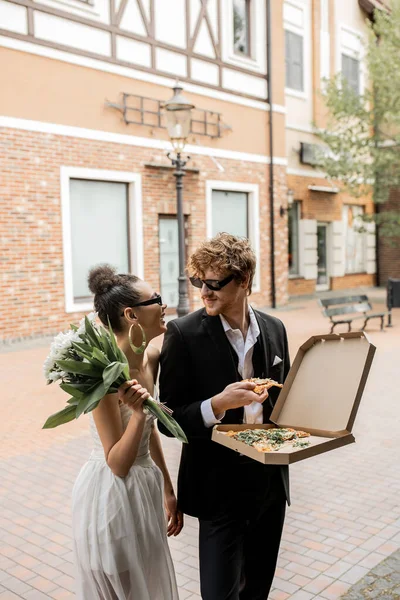 Elegantes recién casados multiétnicos con flores en pizza en la calle urbana, atuendo de boda, gafas de sol - foto de stock