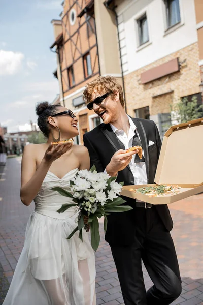 Interracial couple with pizza and flowers smiling at each other in city, sunglasses, wedding outfit — Stock Photo