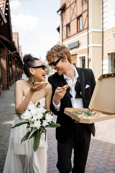 Young multiethnic newlyweds with pizza and flowers laughing on city street, outdoor wedding — Stock Photo