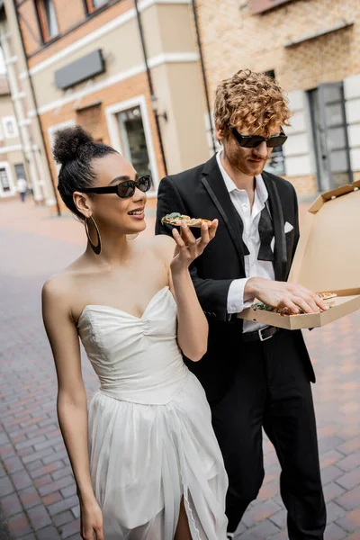 Celebração ao ar livre, casal interracial andando com pizza na cidade, traje de casamento, óculos de sol — Fotografia de Stock