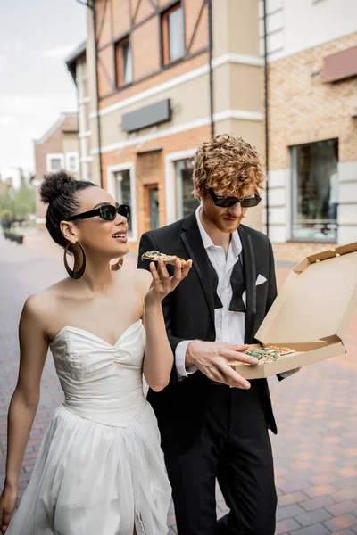 Elegant multiethnic newlyweds in sunglasses walking with pizza in european city, outdoor wedding — Stock Photo