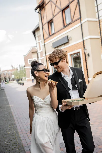 Amour en ville, heureux jeunes mariés multiethniques dans des lunettes de soleil marche avec pizza dans la rue — Photo de stock