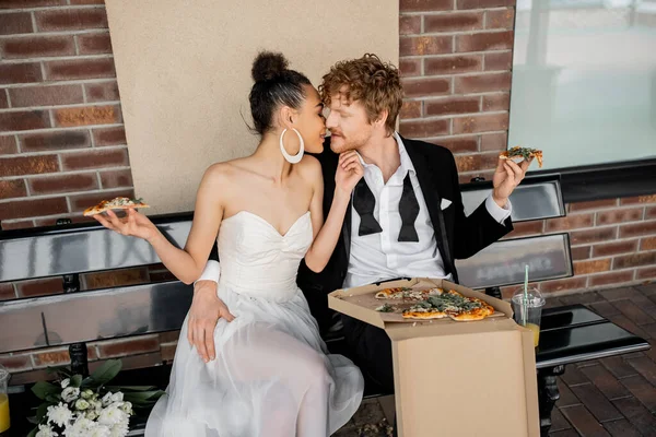 Moderno interracial recién casados con pizza sentado en el banco de la ciudad, celebración al aire libre, felicidad - foto de stock