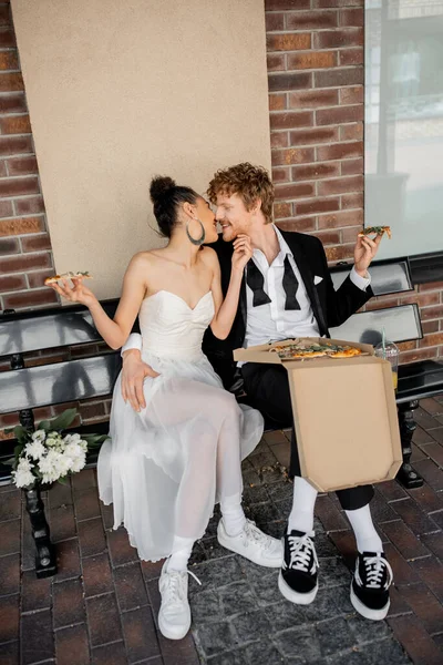 Giovani sposi interrazziale con pizza seduta sulla panchina nella città europea, celebrazione all'aperto — Foto stock