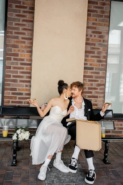 Romantic interracial couple in wedding attire, with pizza, kissing on bench, celebration in city — Stock Photo