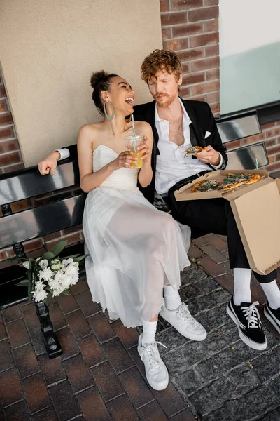 African american bride with orange juice laughing near redhead groom and pizza on bench in city — Stock Photo