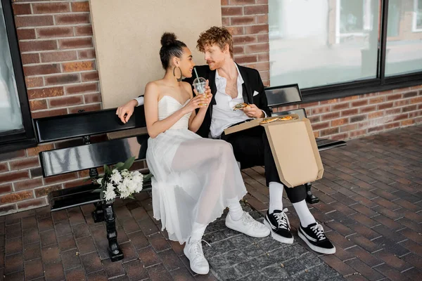 Happy interracial newlyweds with pizza and orange juice looking at each other on bench, city wedding — Stock Photo