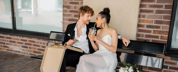Deliziata coppia multietnica appena sposata con pizza e succo d'arancia seduta sulla panchina in città, striscione — Foto stock